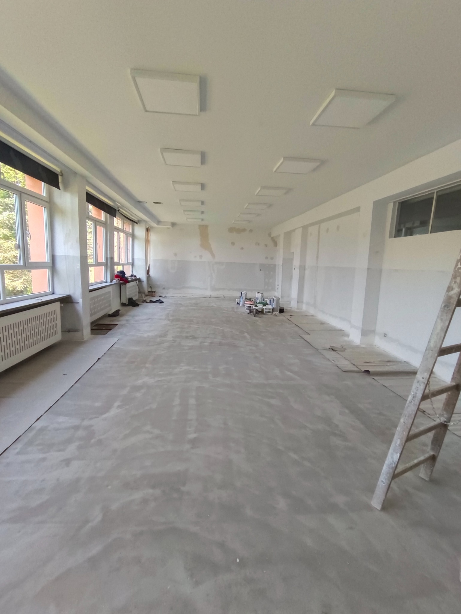 Wnętrze w trakcie prac: gładź na ścianach, wylewka na podłodze, widoczne puszki z farbą i drabina. Duże okna z grzejnikami. Sufit z panelami led.