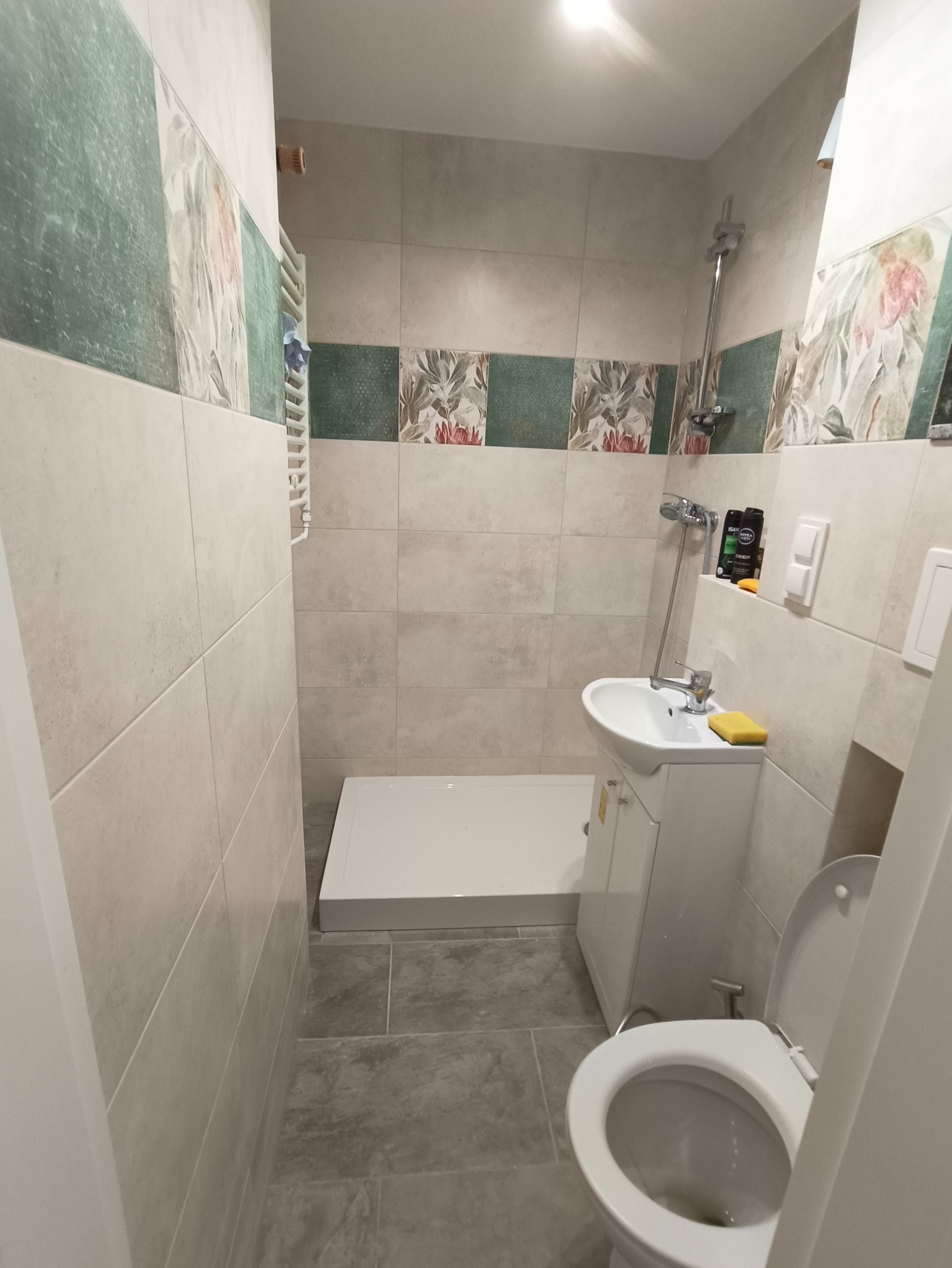 Wykończona łazienka w beżowej tonacji z akcentami kwiatowymi. Kabina prysznicowa z niskim brodzikiem, umywalka z szafką i toaleta. Na ścianach płytki imitujące kamień.