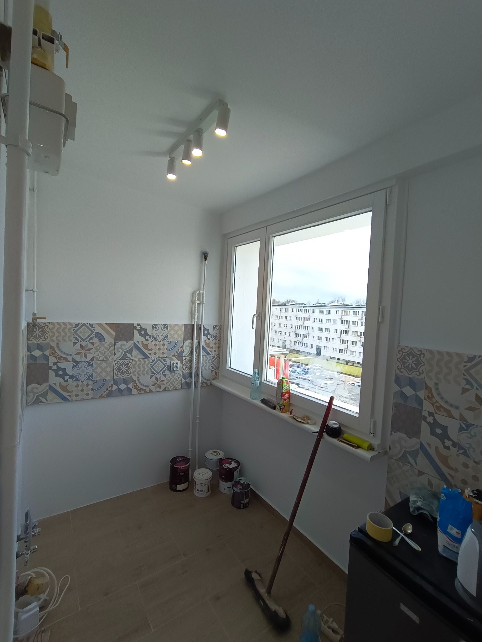 Wnętrze w trakcie prac wykończeniowych: pomalowane ściany, ułożone płytki dekoracyjne, widoczne puszki z farbą, okno z widokiem na blok.