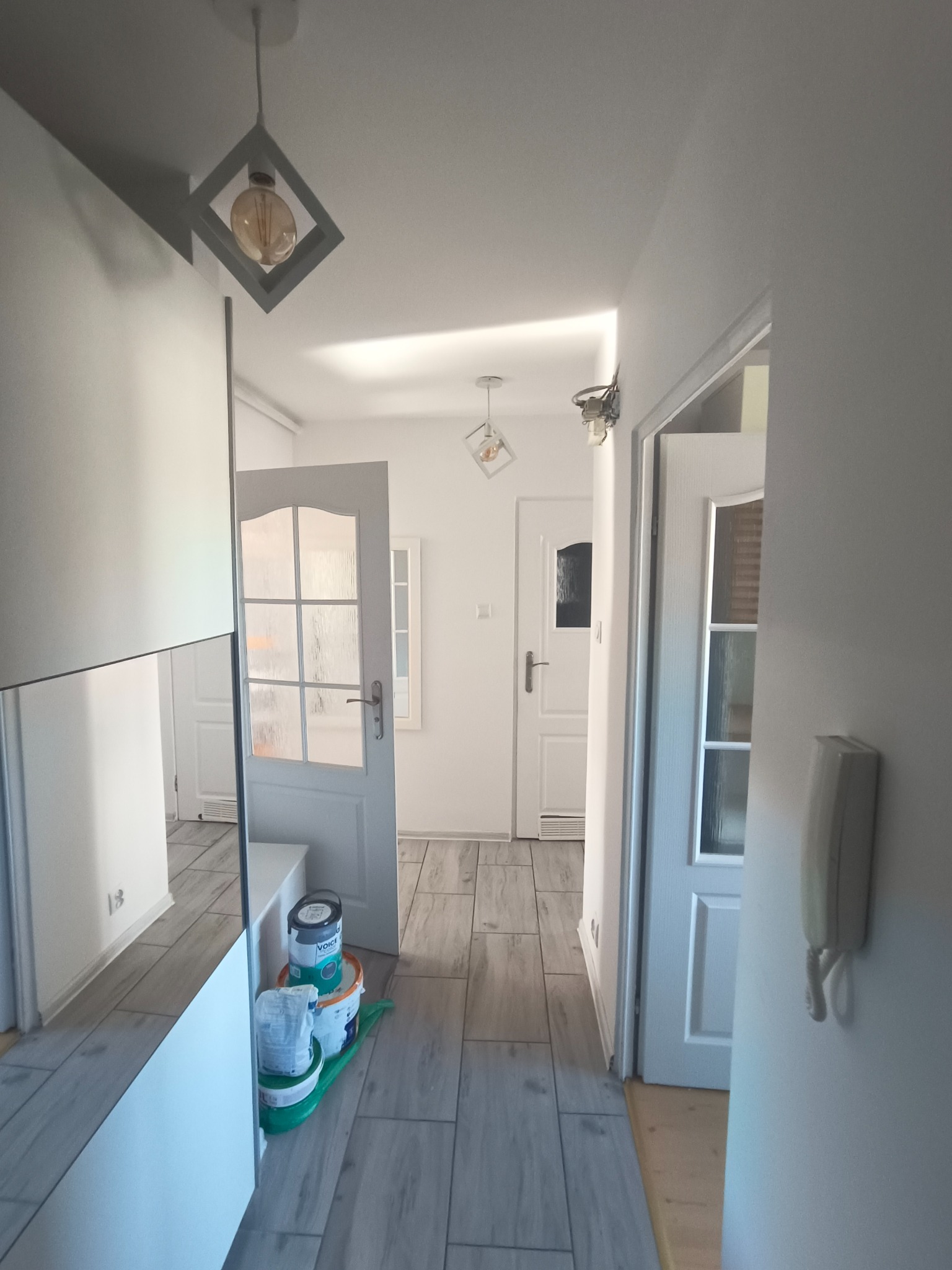 Jasne wnętrze korytarza po remoncie z szarą podłogą imitującą drewno, widoczne puszki z farbą i nowoczesne oświetlenie. Otwarta przestrzeń z nowymi drzwiami.