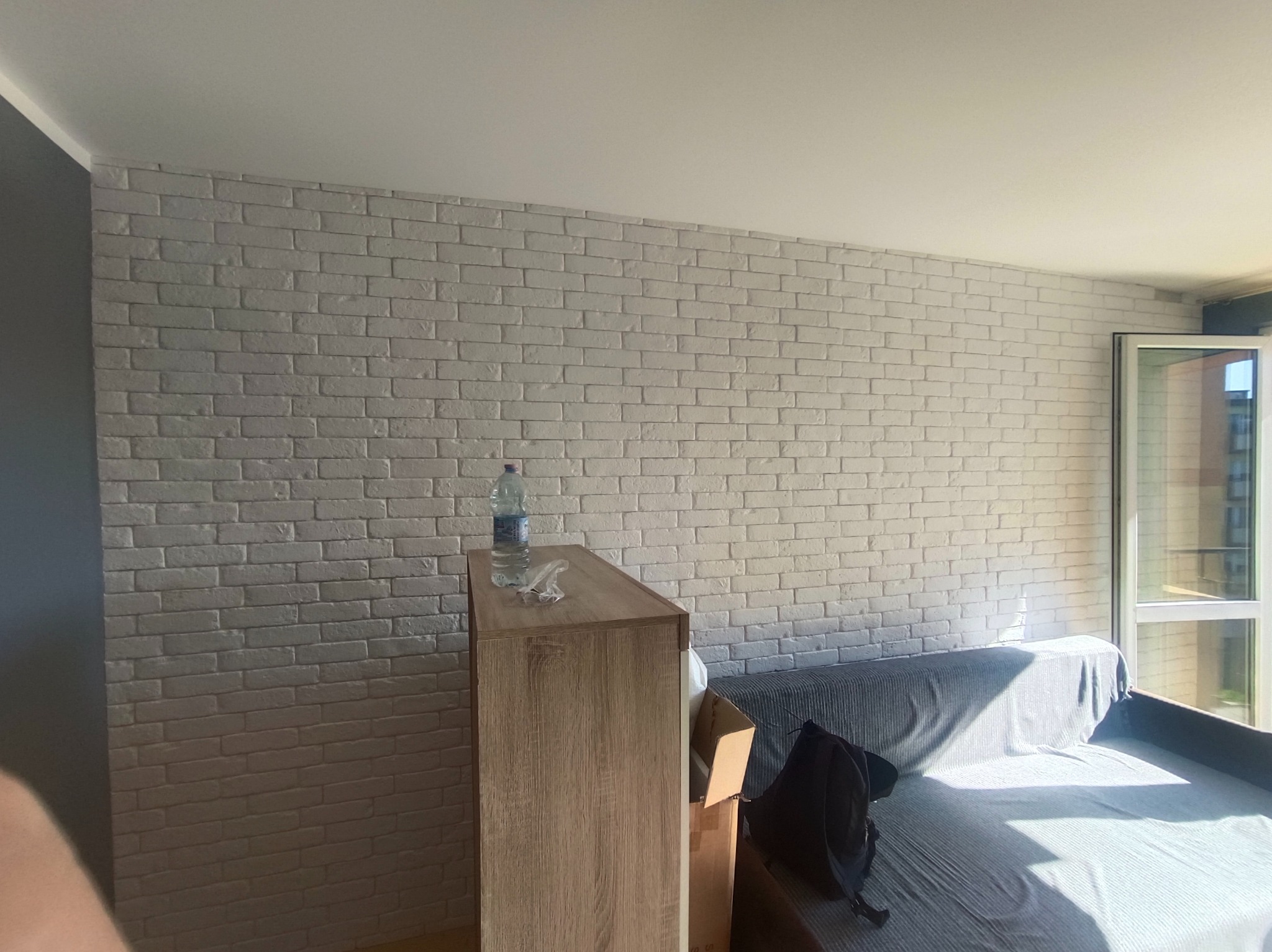 Ściana z białej cegły dekoracyjnej w pokoju, widoczna część sofy i szafki. Minimalistyczne wnętrze z elementami dekoracyjnymi, jasne oświetlenie.