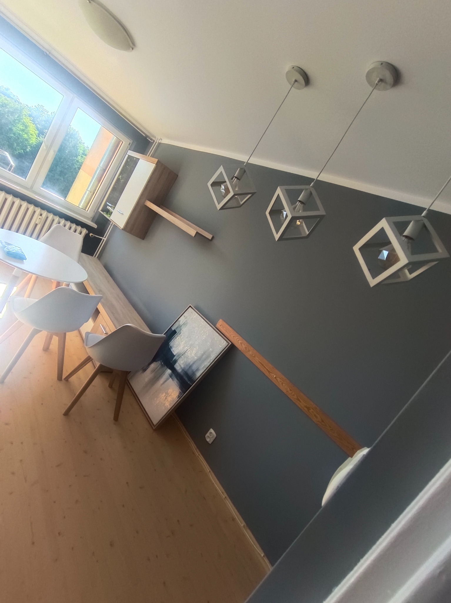 Nowoczesne wnętrze: drewniana podłoga, szare ściany, geometryczne lampy wiszące, stół z krzesłami i obraz oparty o ścianę. Nietypowy kąt ujęcia.