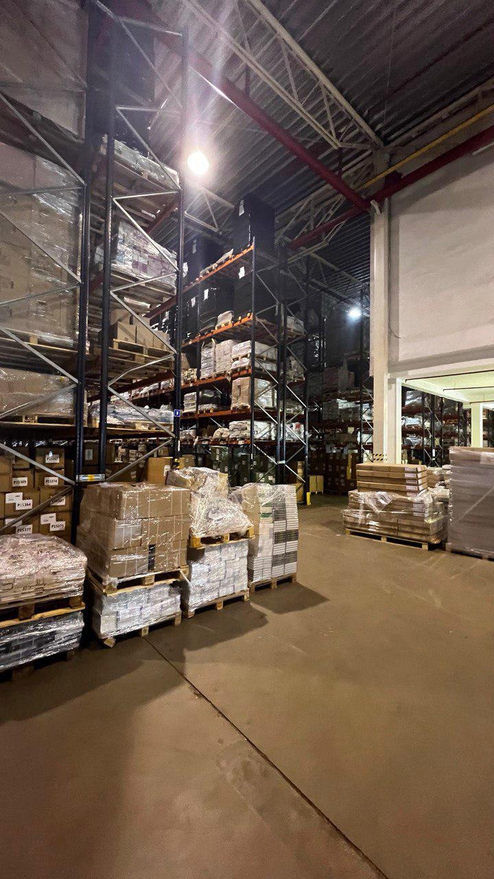 Magazyn z wysokimi regałami wypełnionymi paletami z towarem, niektóre owinięte folią stretch, oświetlony jasnym światłem z góry.