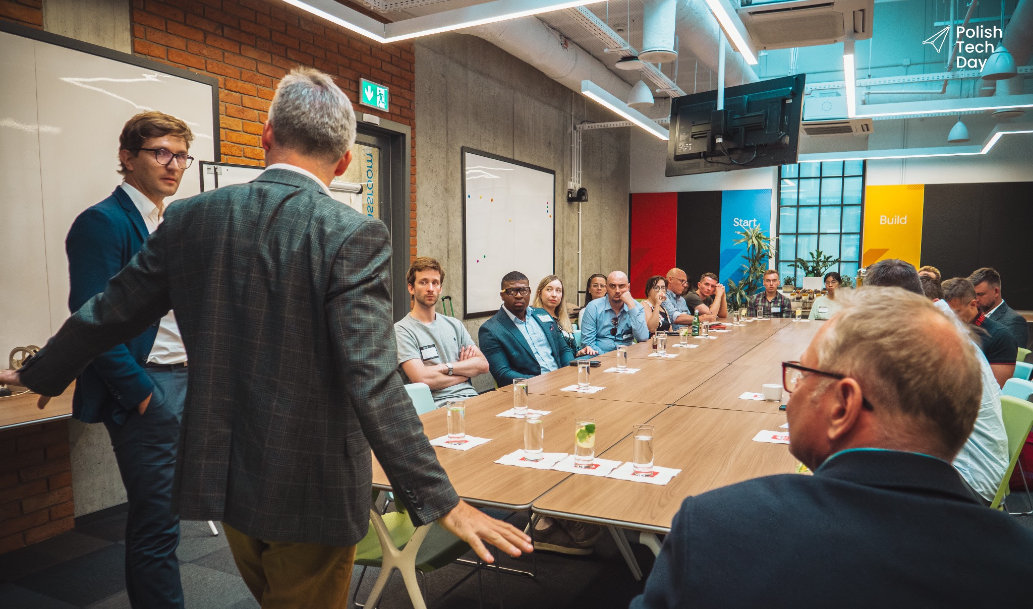 Sala konferencyjna podczas Polish Tech Day: Grupa osób siedzi przy długim stole, słuchając prezentacji; widoczne szklanki z wodą i logo wydarzenia w tle.