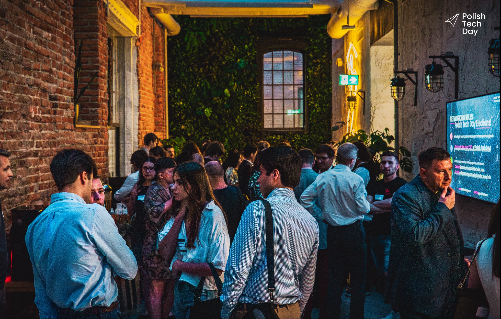 Fotorelacja z wydarzenia Polish Tech Day: tłum ludzi w przestrzeni z ceglaną ścianą, ścianą z roślin, oświetleniem neonowym i ekranem wyświetlającym zasady networkingu.