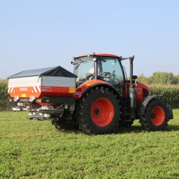 P.H. Agroma S.A. w Poznaniu - Czerwony ciągnik Kubota z rozsiewaczem nawozów w trakcie pracy na polu, w tle widoczne wysokie kukurydze i błękitne niebo.