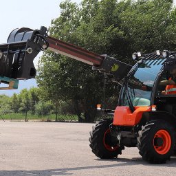 P.H. Agroma S.A. w Poznaniu - Pomarańczowa ładowarka teleskopowa Taurulift podnosi oponę na palecie, operator w kamizelce odblaskowej w kabinie, w tle zielone drzewa i fragment boiska.