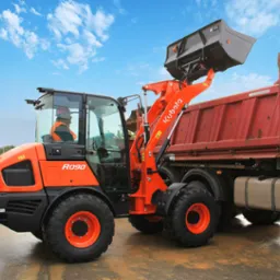 Pomarańczowa miniładowarka Kubota R090 z podniesioną łyżką ładuje czerwoną wywrotkę na tle błękitnego nieba z chmurami.