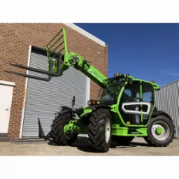 Zielona ładowarka teleskopowa Merlo TF35.7 na tle ceglanego budynku z zamkniętą bramą rolowaną, widok z boku, czyste niebo w tle.