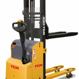 Żółto-szary wózek paletowy elektryczny marki TCM na białym tle, widok z boku. Widoczna konstrukcja masztu z łańcuchem i zabezpieczeniem, dyszel sterujący i palety.
