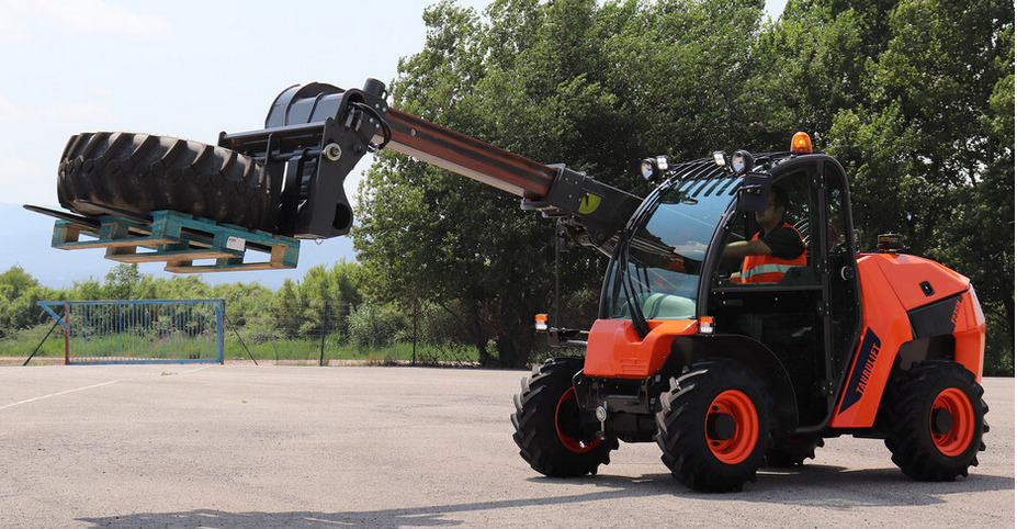Pomarańczowa ładowarka teleskopowa Taurulift podnosi oponę na palecie, operator w kamizelce odblaskowej w kabinie, w tle zielone drzewa i fragment boiska.