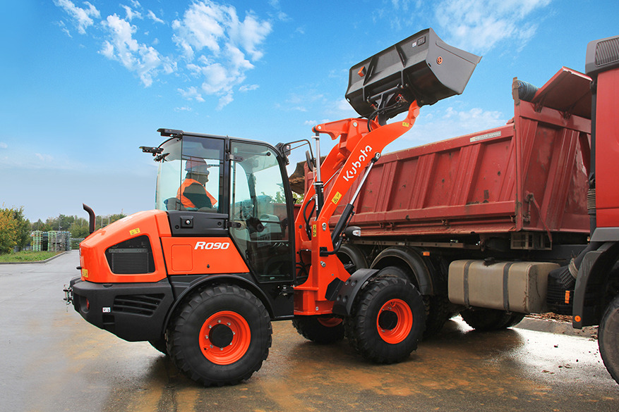 Pomarańczowa miniładowarka Kubota R090 z podniesioną łyżką ładuje czerwoną wywrotkę na tle błękitnego nieba z chmurami.
