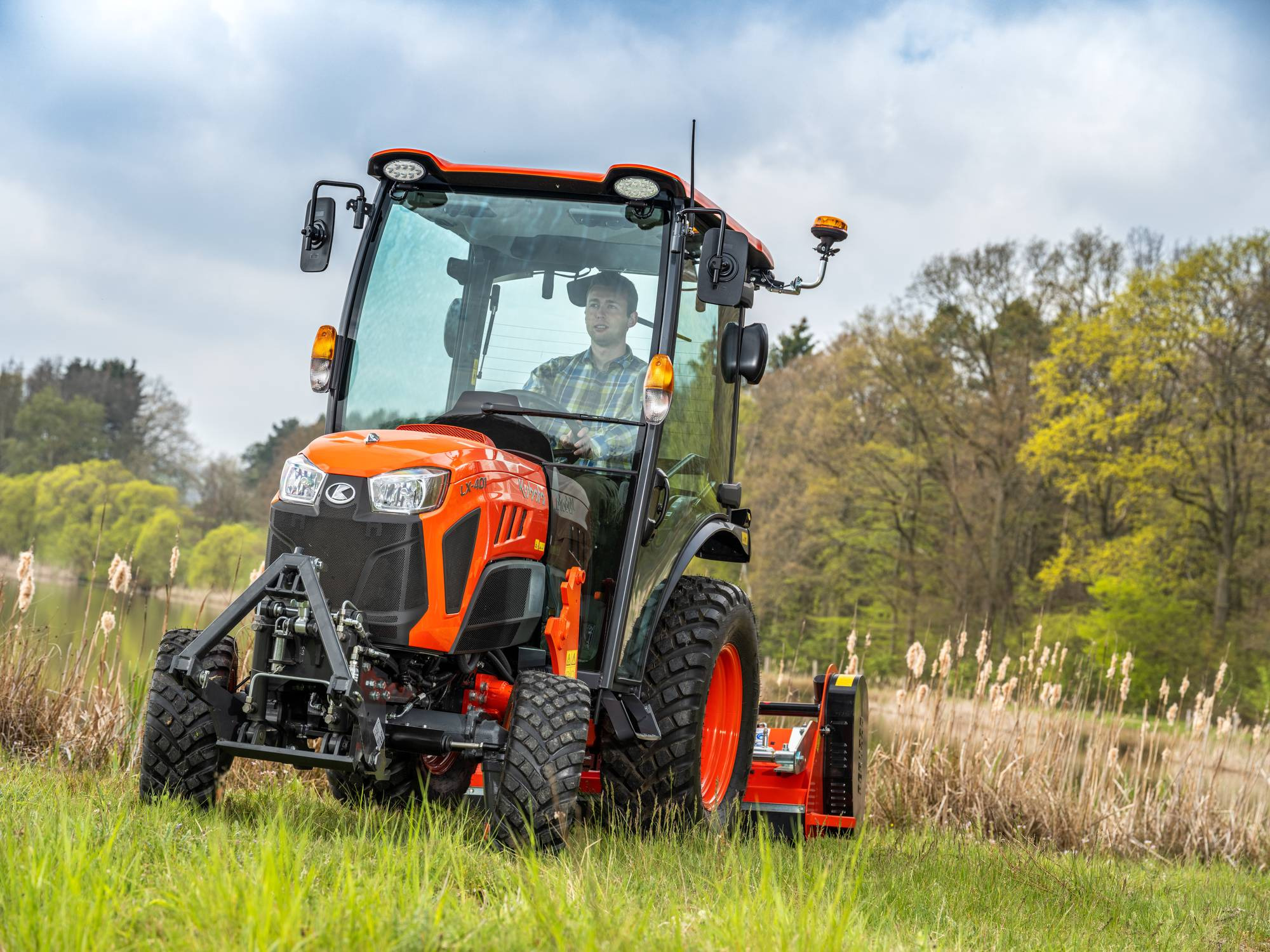 Pomarańczowy ciągnik Kubota LX-401 z kabiną, prowadzony przez mężczyznę w kraciastej koszuli, na tle jeziora i drzew.