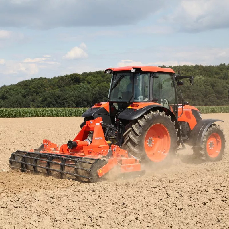 Pomarańczowy traktor marki Kubota z glebogryzarką w trakcie pracy na polu, widok z boku, zalesione wzgórze w tle.