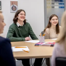 Uczniowie uśmiechnięci podczas zajęć w jasnym pomieszczeniu, na stole leżą zeszyty i przybory do pisania, na ścianie wiszą obrazy.
