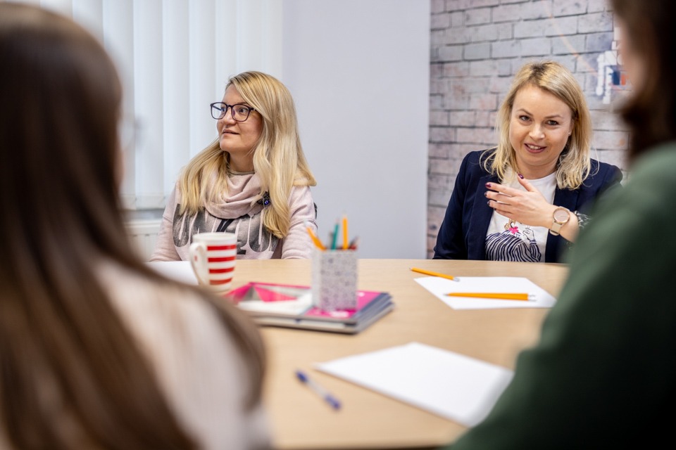 Dwie kobiety siedzące przy stole podczas lekcji angielskiego, jedna z nich z blond włosami i okularami spogląda w bok, druga w granatowej marynarce gestykuluje podczas rozmowy, na stole leżą...