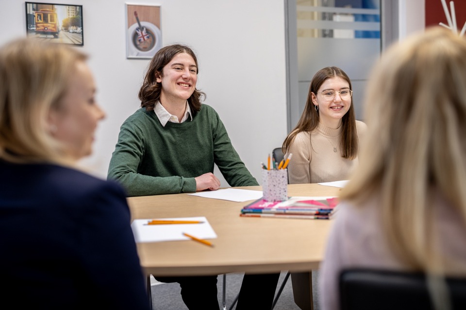 Uczniowie uśmiechnięci podczas zajęć w jasnym pomieszczeniu, na stole leżą zeszyty i przybory do pisania, na ścianie wiszą obrazy.