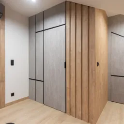 Nowoczesne wnętrze z zabudową w odcieniach szarości i drewna, obejmujące szafy z minimalistycznymi uchwytami i półki w nietypowym kształcie, uzupełnione drewnianymi panelami ściennymi...