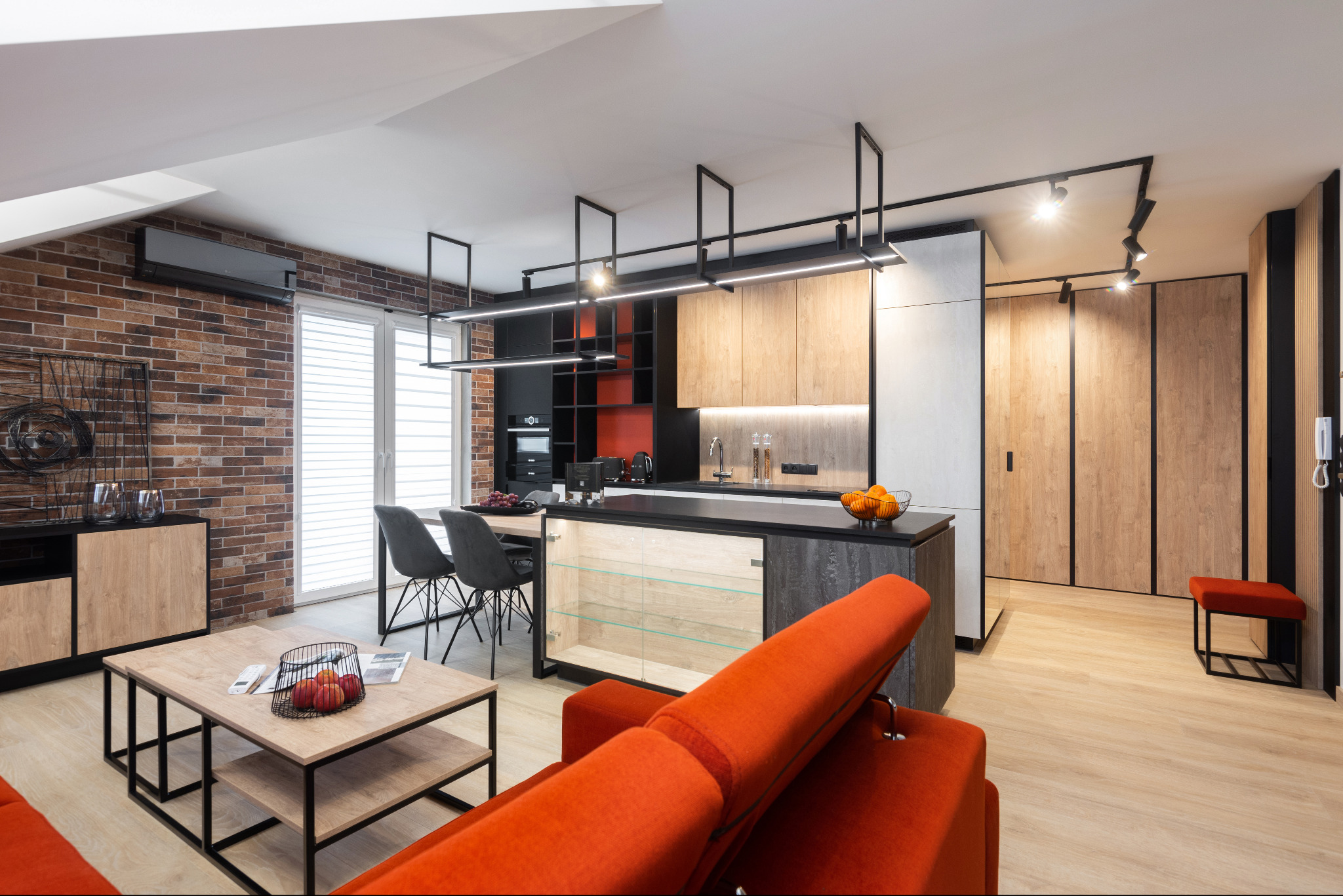 Nowoczesne wnętrze mieszkania z otwartą kuchnią, ceglaną ścianą, wyspą kuchenną z przeszklonymi drzwiczkami, pomarańczową sofą i designerskim oświetleniem.