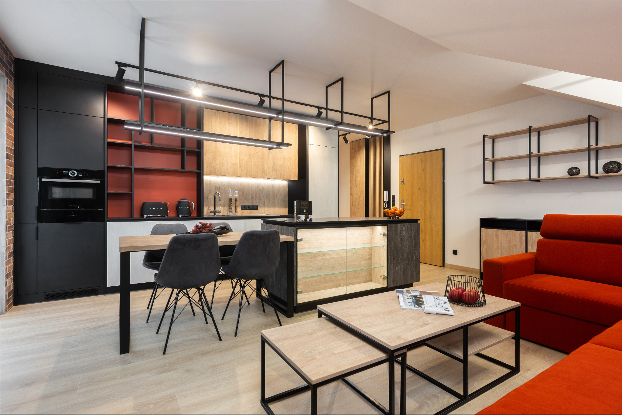 Nowoczesne wnętrze z czarną zabudową kuchenną, drewnianymi akcentami, oświetleniem LED i czerwonymi elementami dekoracyjnymi, w tym sofą i akcesoriami kuchennymi.