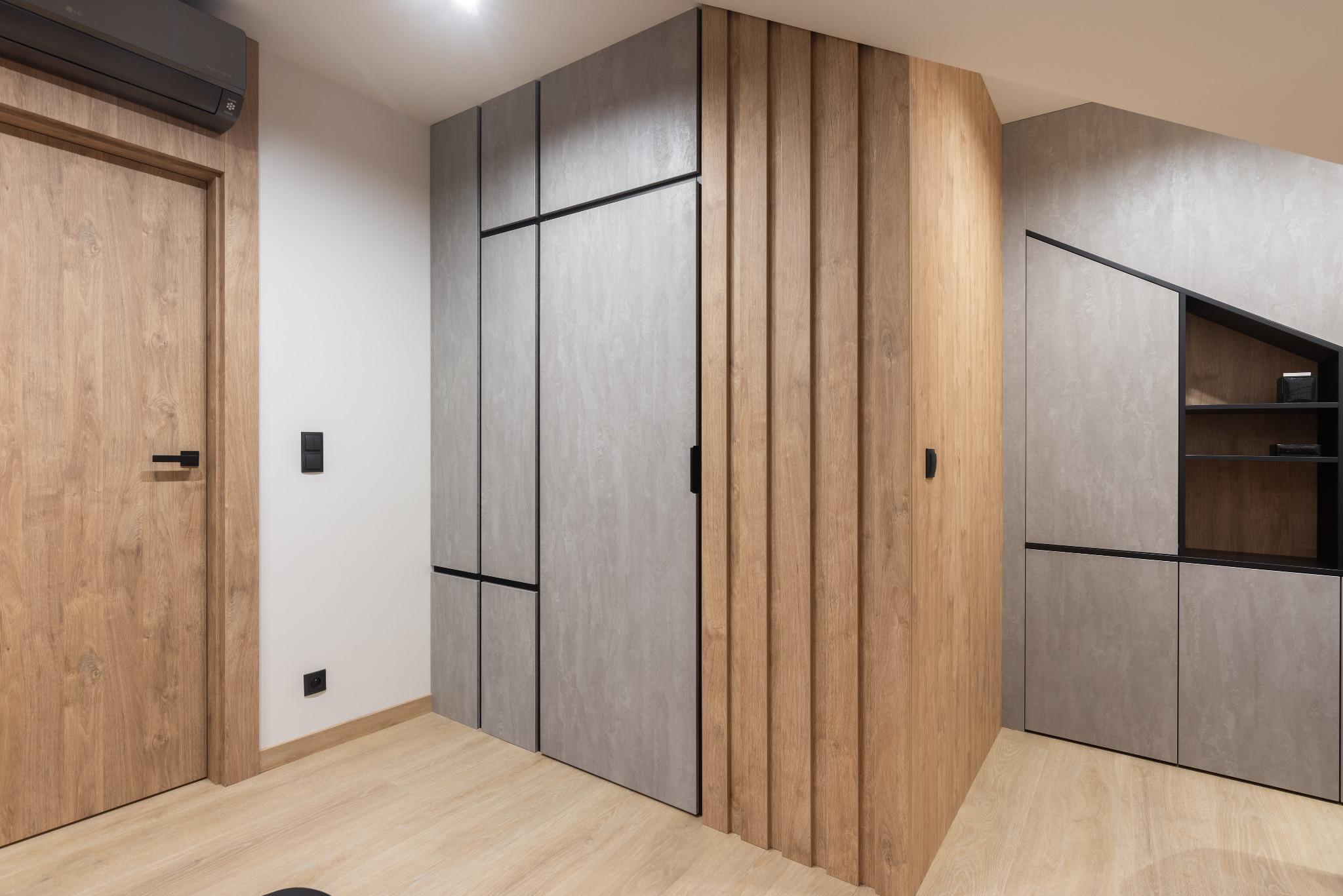 Nowoczesne wnętrze z zabudową w odcieniach szarości i drewna, obejmujące szafy z minimalistycznymi uchwytami i półki w nietypowym kształcie, uzupełnione drewnianymi panelami ściennymi...