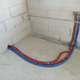 Wewnętrzne instalacje hydrauliczne i grzewcze w surowym stanie, rury prowadzone wzdłuż ścian z betonu komórkowego, zabezpieczone opaskami, widoczne otwory w ścianie i podłodze.