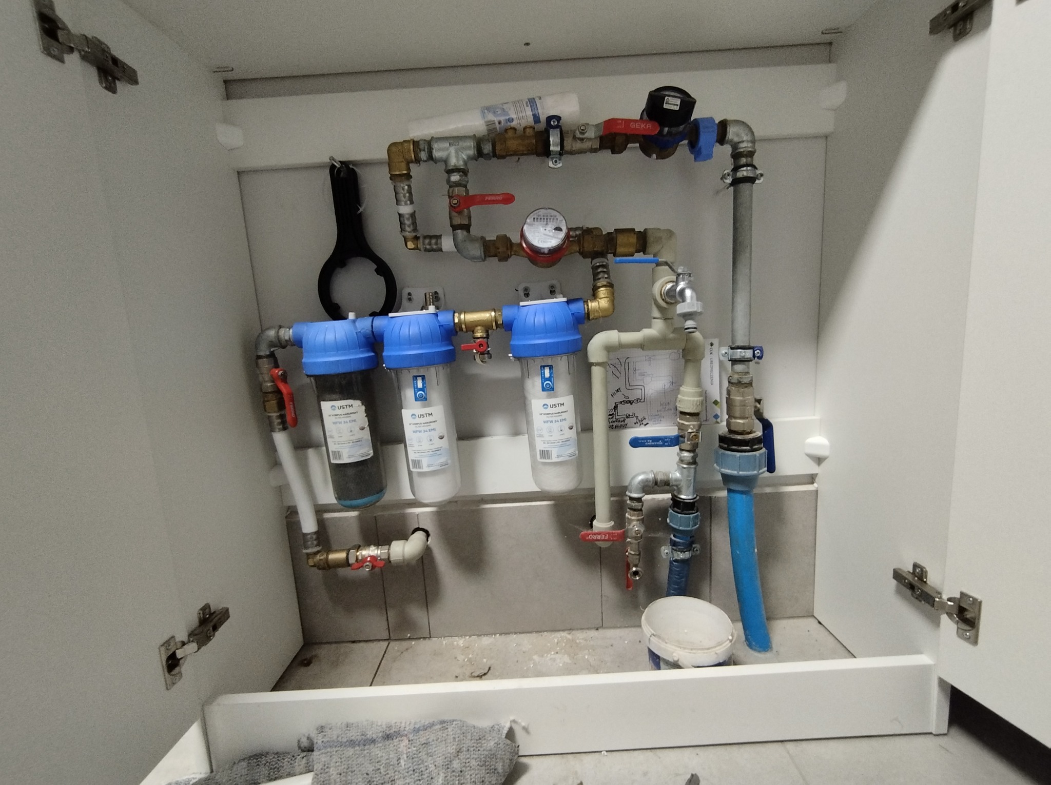 Instalacja wodna z filtrami mechanicznymi i licznikiem, umieszczona w szafce pod zlewem, widoczne rury miedziane i plastikowe, zawory kulowe z czerwonymi rączkami, klucz do filtra zawieszony...