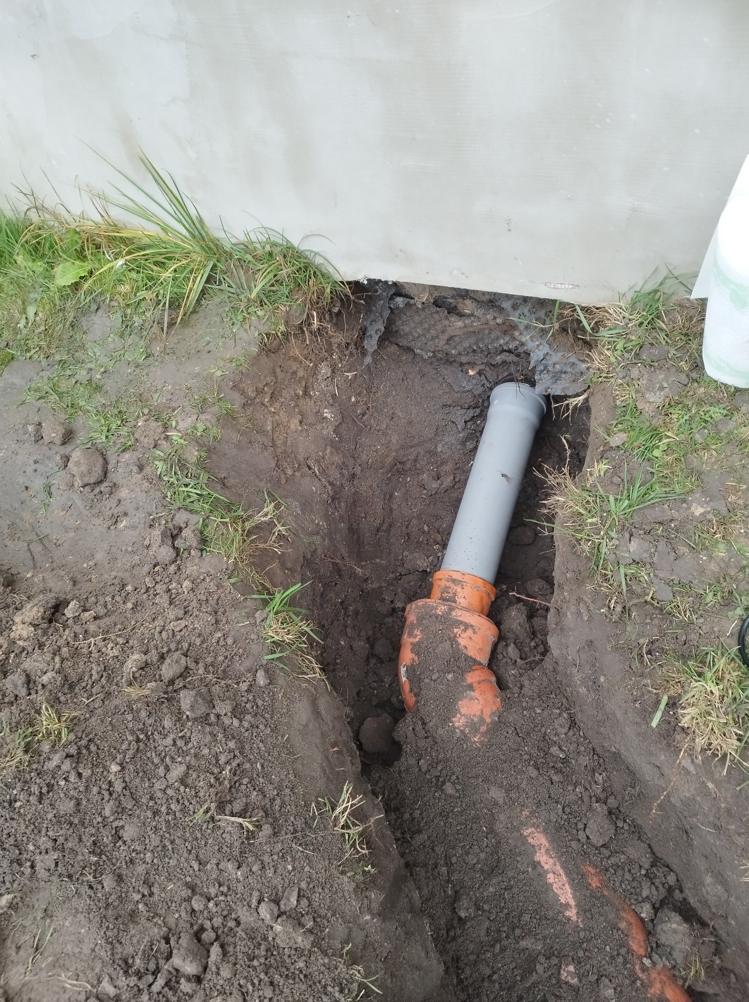 Fragment instalacji kanalizacyjnej z szarej i pomarańczowej rury w wykopie ziemnym, widoczne połączenie rur i fragment ściany budynku w tle.