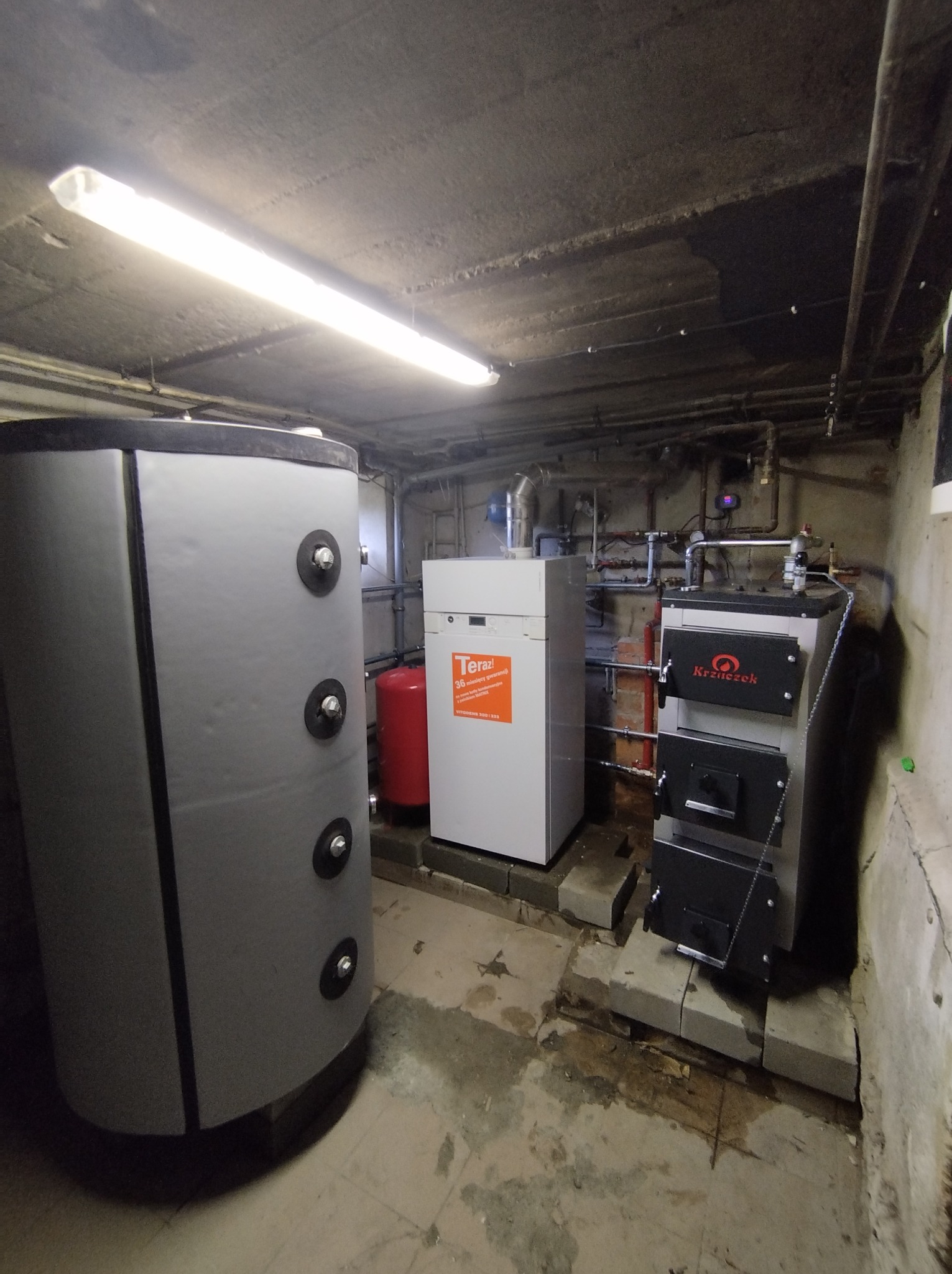 Kotłownia z trzema różnymi piecami: zasobnikiem wody, piecem gazowym z naklejką i piecem na paliwo stałe, widać liczne rury i instalacje na ścianie.