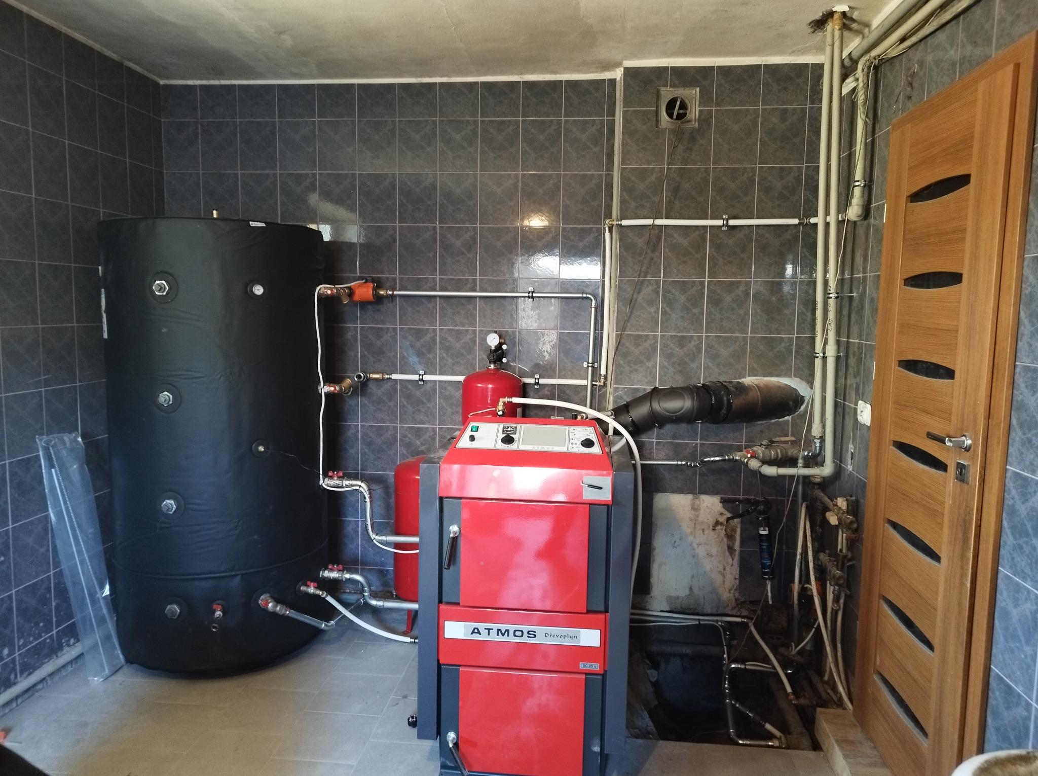 Czerwony piec marki Atmos Drevo plyn z podłączonymi rurami i czarnym zasobnikiem na wodę w pomieszczeniu z szarymi płytkami.