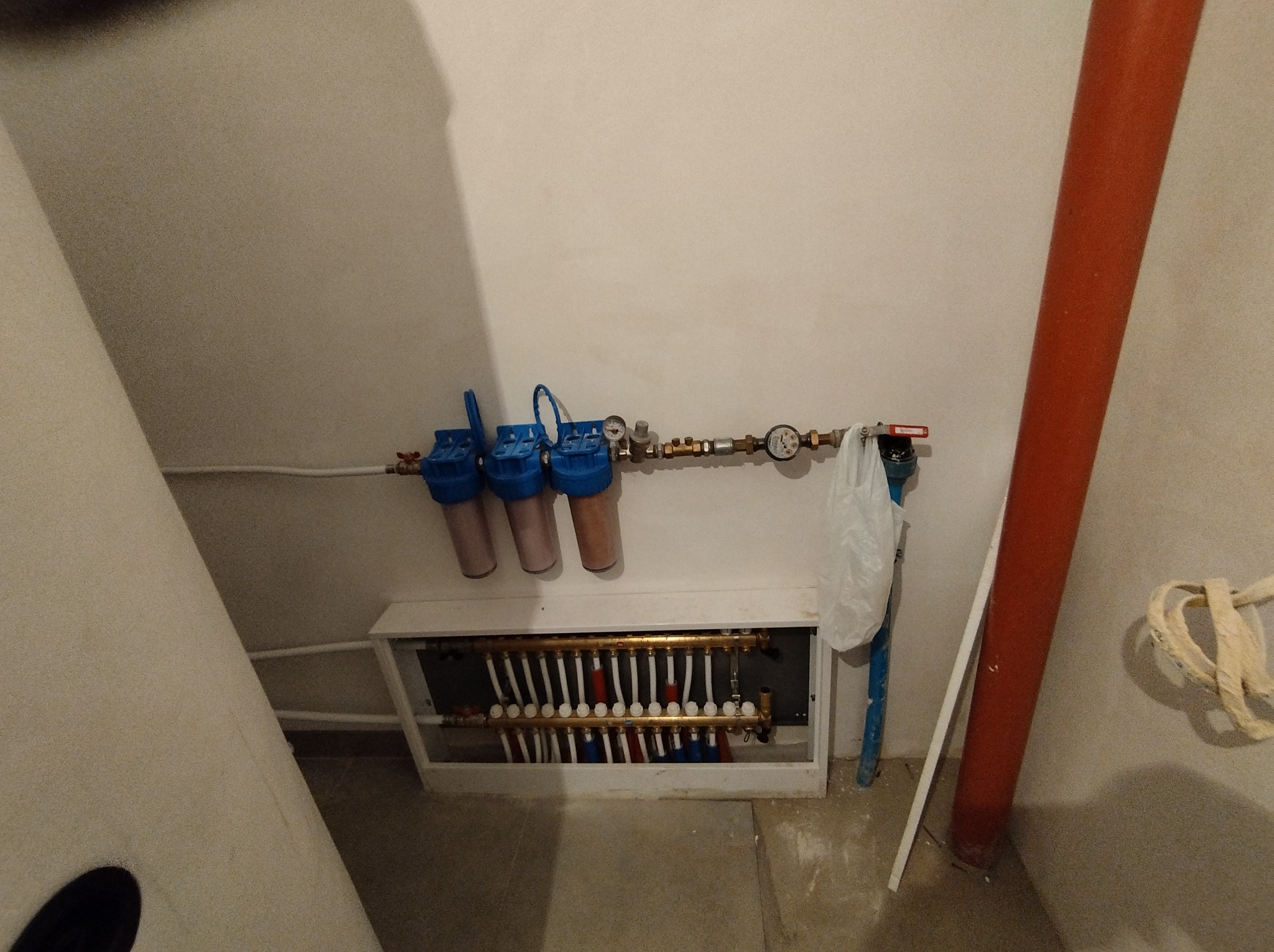 Rozdzielacz ogrzewania podłogowego z filtrami wody i manometrem, zamontowany w szafce pod tynkiem, widoczna rura pionowa w kolorze czerwonym.