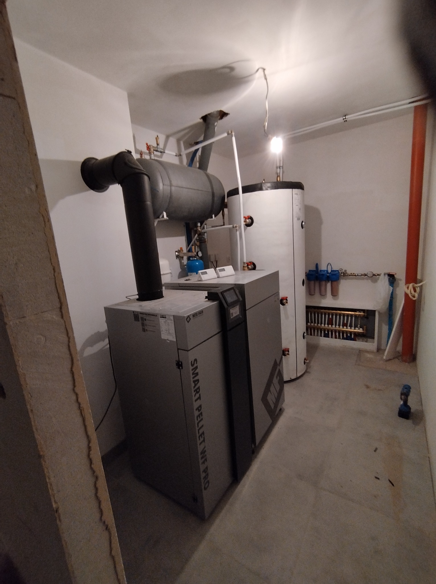 Szary piec na pellet Smart Pellet WF PRO z podłączonym kominem, obok biały zasobnik ciepłej wody i rozdzielacz ogrzewania podłogowego w kotłowni, widoczny betonowy słup konstrukcyjny.