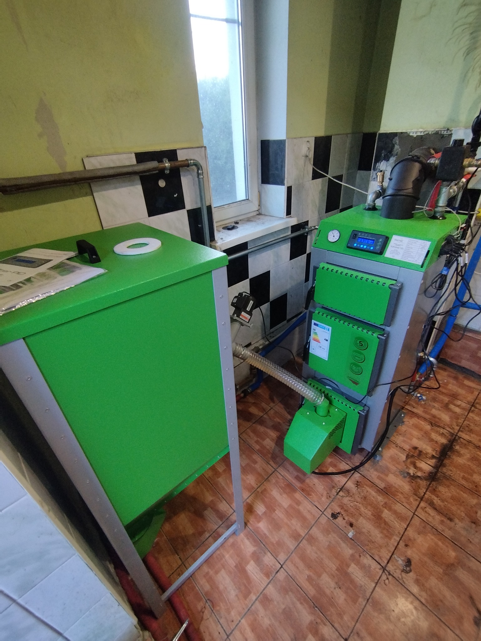 Nowoczesny, zielono-szary piec CO z podajnikiem na ekogroszek, zainstalowany w pomieszczeniu z kafelkami na ścianach i podłodze, widoczny także zasobnik na paliwo.