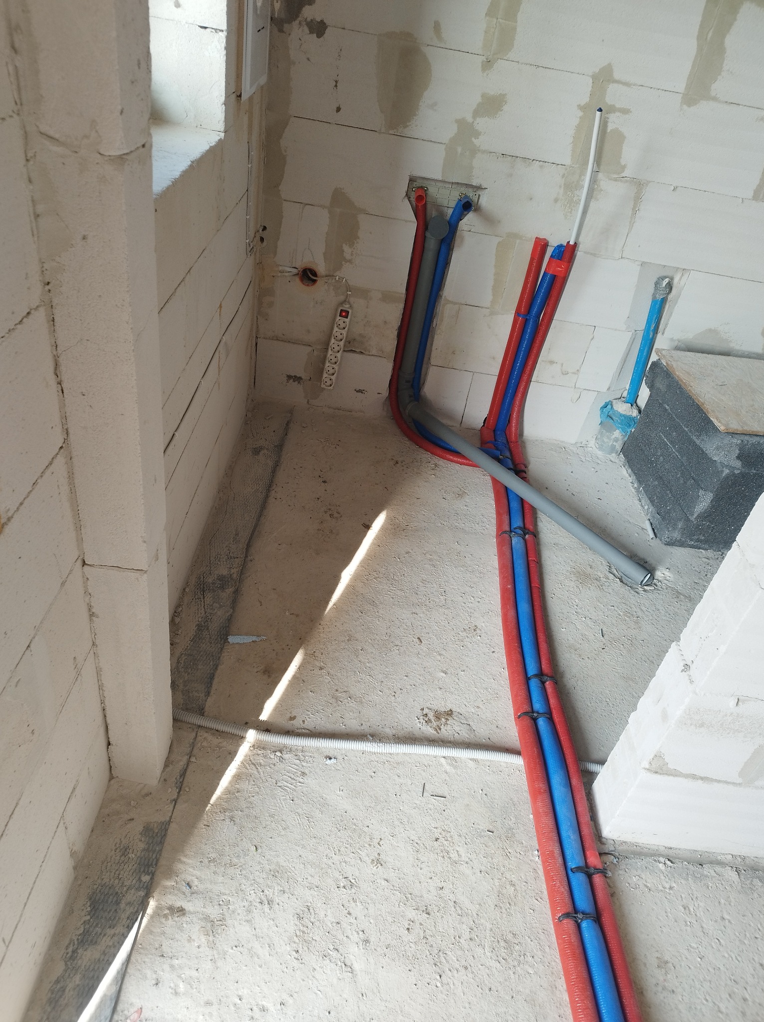 Surowe wnętrze w trakcie prac instalacyjnych: rury grzewcze (czerwone i niebieskie) oraz szare rury wodne prowadzone wzdłuż ściany z betonu komórkowego, widoczna listwa zasilająca i fragment okna.
