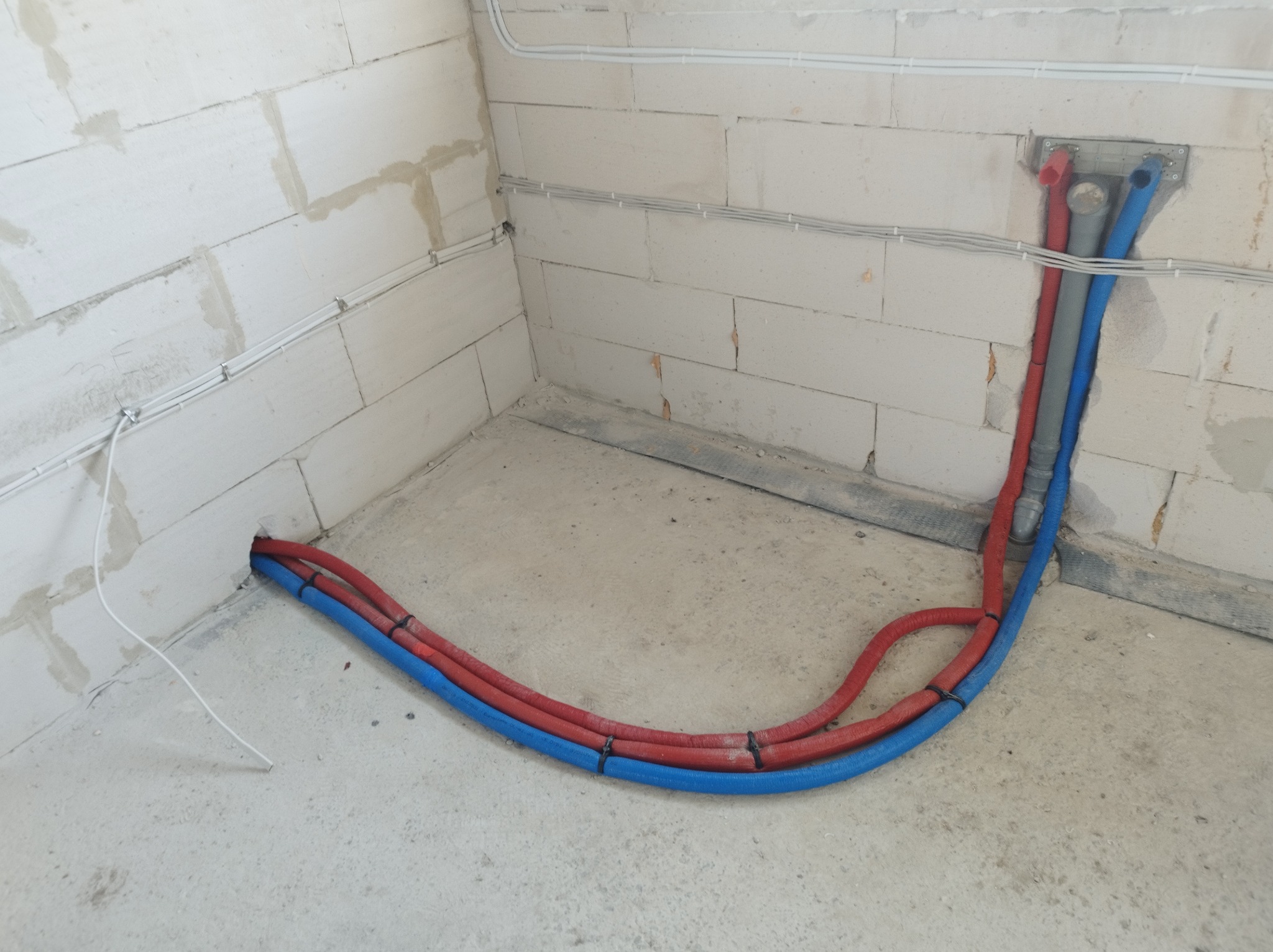 Wewnętrzne instalacje hydrauliczne i grzewcze w surowym stanie, rury prowadzone wzdłuż ścian z betonu komórkowego, zabezpieczone opaskami, widoczne otwory w ścianie i podłodze.