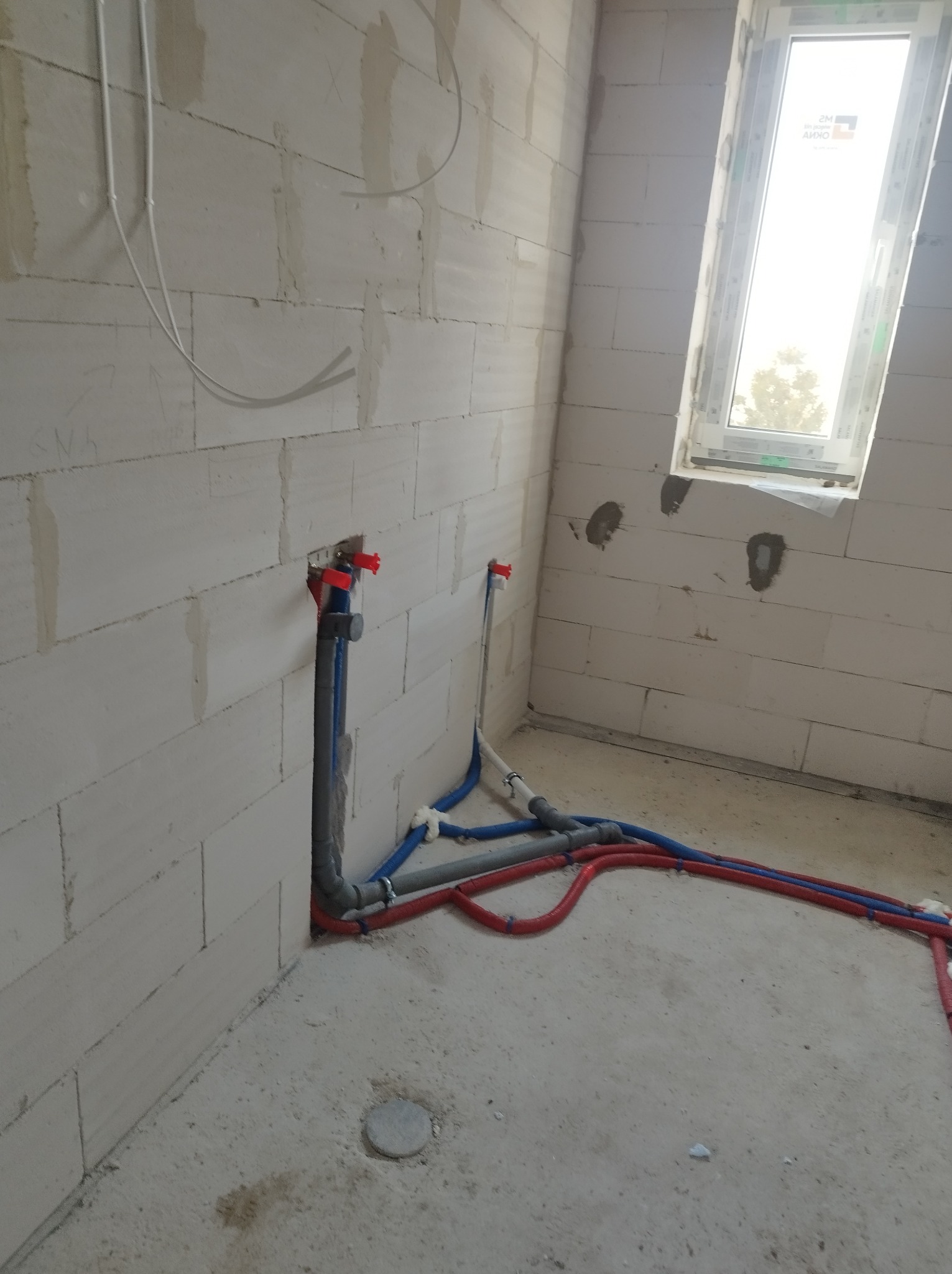Surowy stan instalacji wodnej i grzewczej w pomieszczeniu z bloczków, widoczne rury w kolorach niebieskim i czerwonym, szare rury odpływowe, ściany w trakcie budowy, okno dachowe.