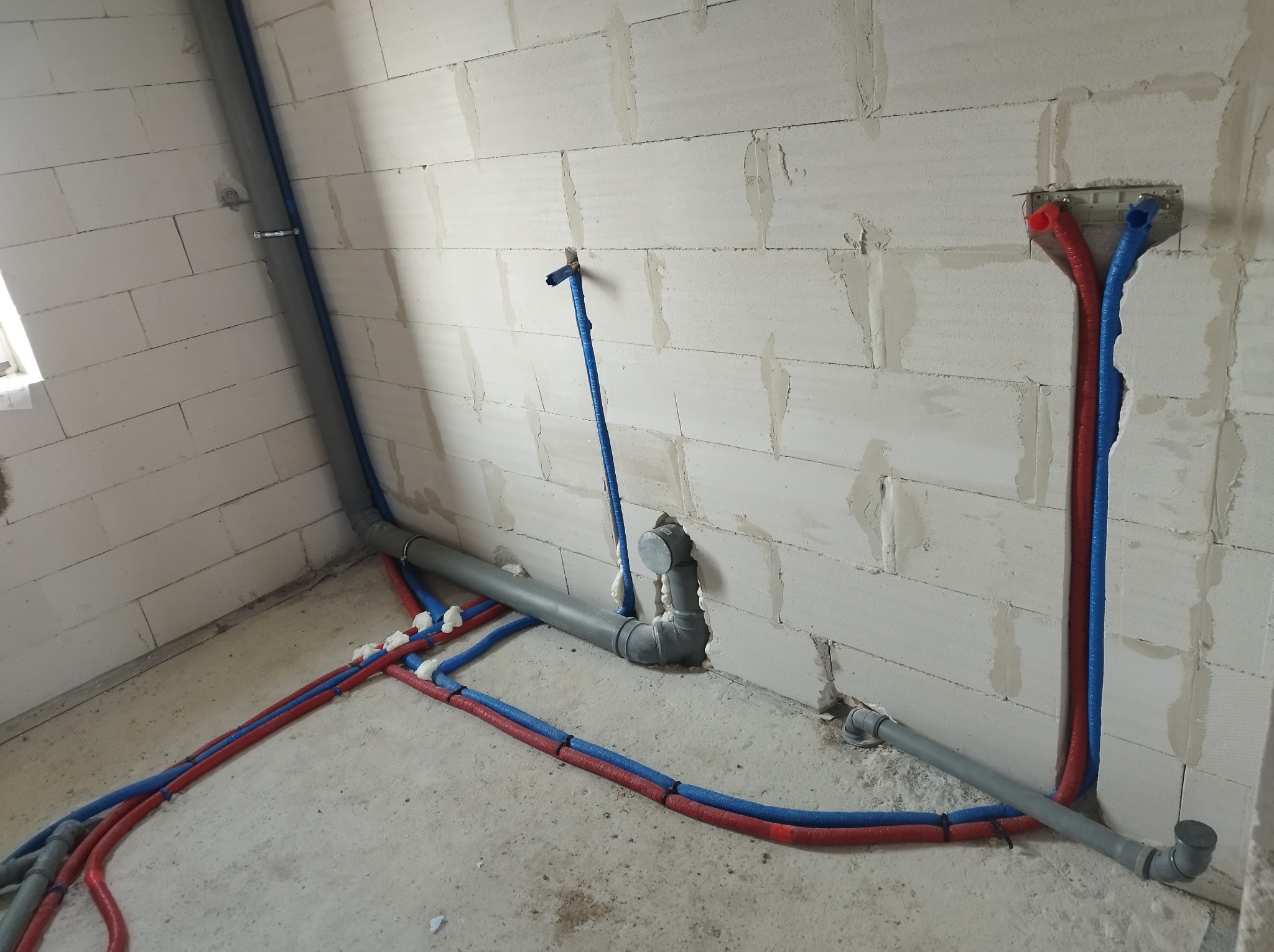 Wewnętrzne instalacje wodne i kanalizacyjne w surowym stanie, rury PEX w izolacji czerwonej i niebieskiej, szare rury kanalizacyjne, ściany z betonu komórkowego.