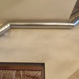 Metalowa rura wentylacyjna zamontowana pod sufitem w rogu pomieszczenia, widoczna osłonięta folią framuga okienna.