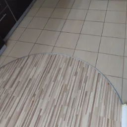 Połączenie dwóch rodzajów podłóg: jasne płytki kwadratowe i panele podłogowe imitujące drewno, oddzielone metalową listwą w kształcie łuku, widoczne szafki kuchenne.