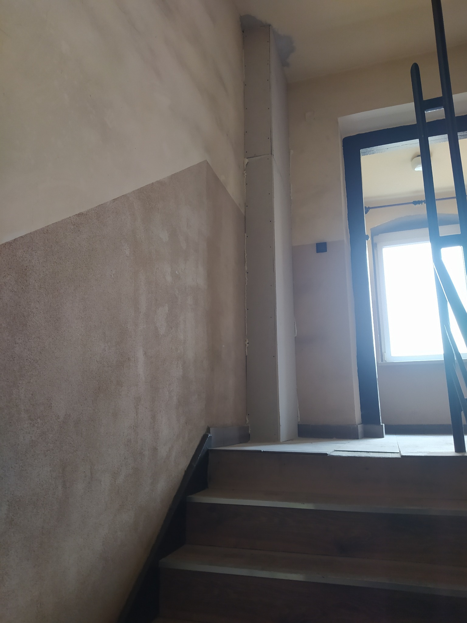 Klatka schodowa w trakcie prac wykończeniowych: ściany z nałożoną strukturą, częściowo obudowany filar karton-gipsem, drewniane schody.