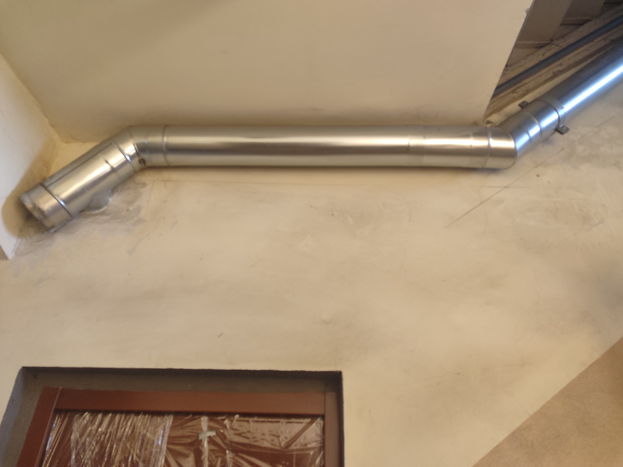 Metalowa rura wentylacyjna zamontowana pod sufitem w rogu pomieszczenia, widoczna osłonięta folią framuga okienna.