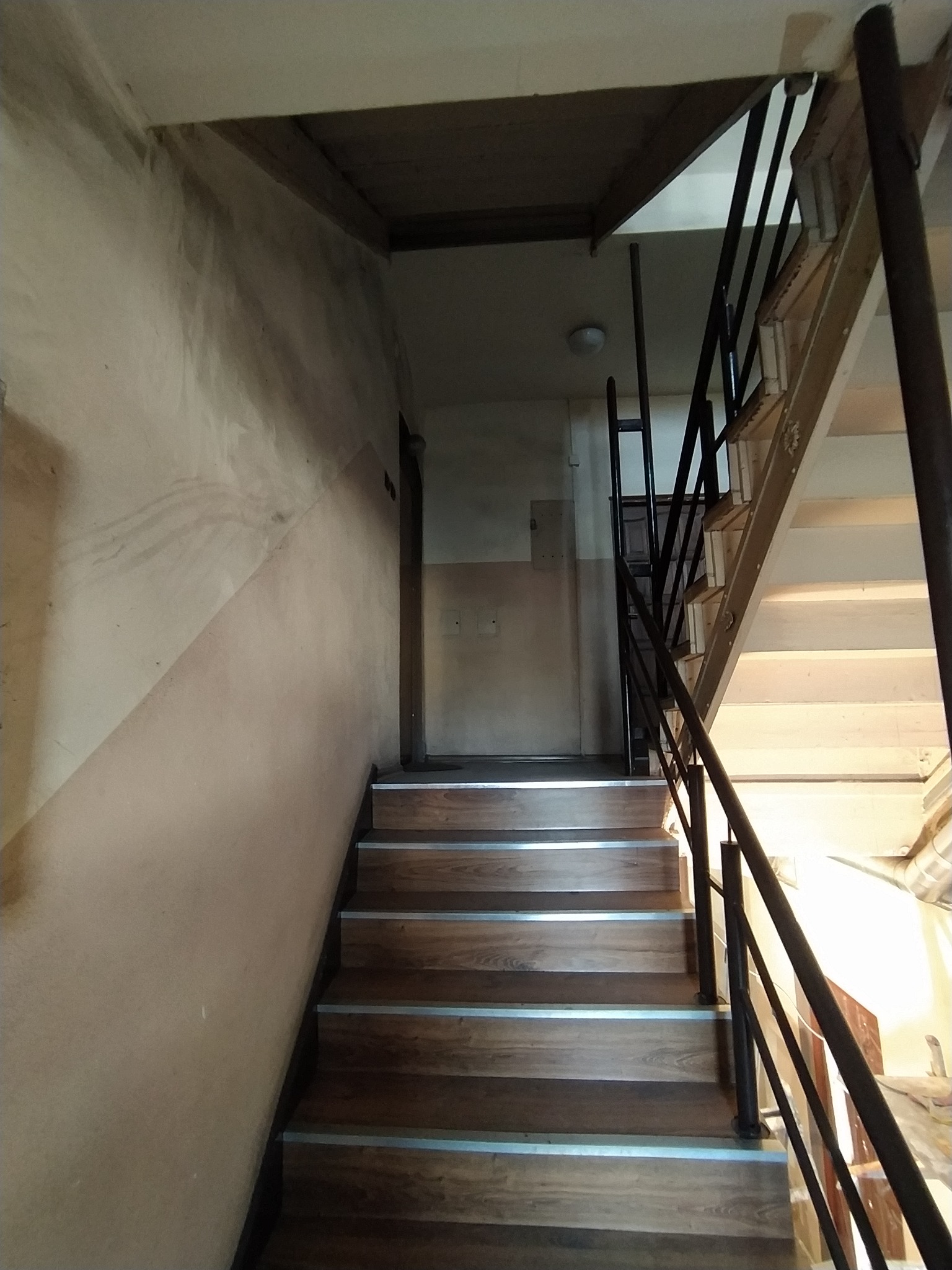 Klatka schodowa z drewnianymi schodami i metalową balustradą, ściany z widocznymi śladami po wcześniejszych pracach malarskich, widoczne różne odcienie farby.