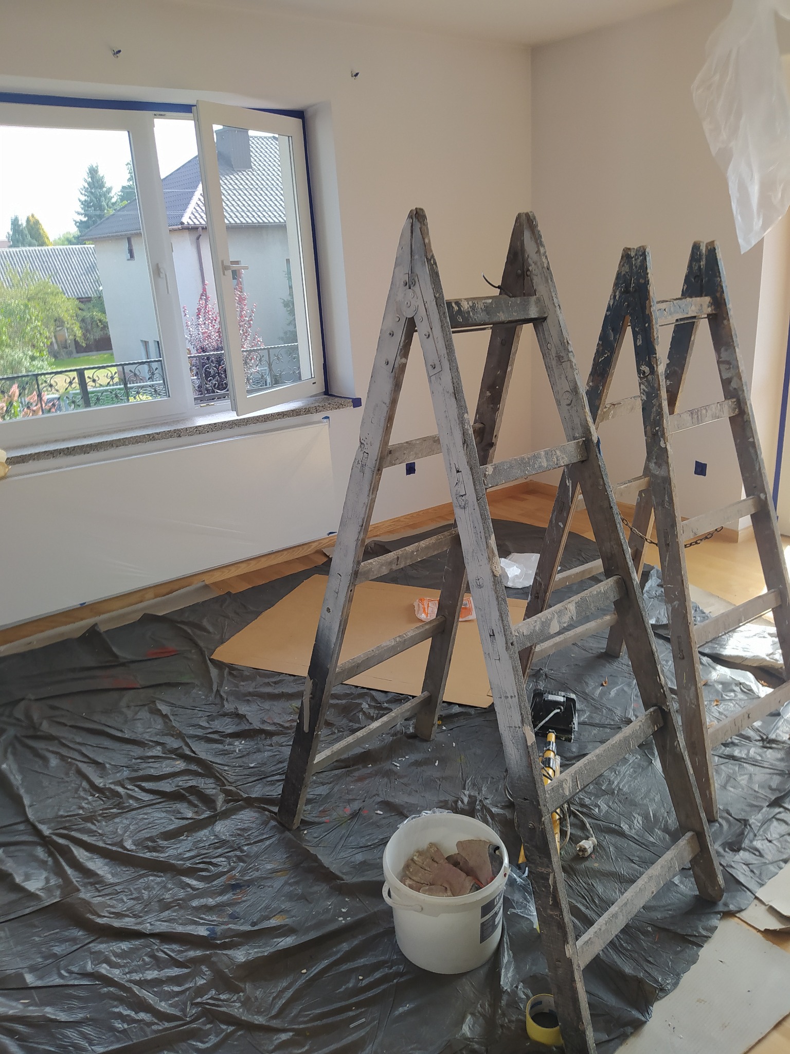 Wnętrze pokoju w trakcie odświeżania: drewniane schody malarskie, podłoga zabezpieczona folią, okno z taśmą malarską, wiadro z rękawicami malarskimi.