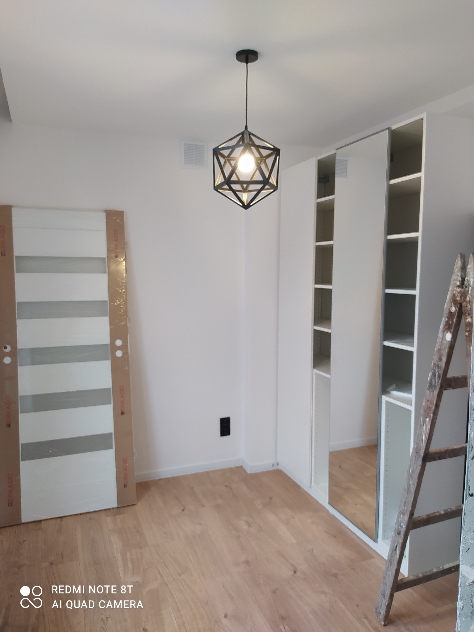 Wnętrze pokoju w trakcie wykańczania: zamontowana biała szafa z lustrem, podłoga z paneli, drzwi z szybami w kartonowym opakowaniu oparte o ścianę, metalowa drabina i nowoczesna lampa sufitowa.