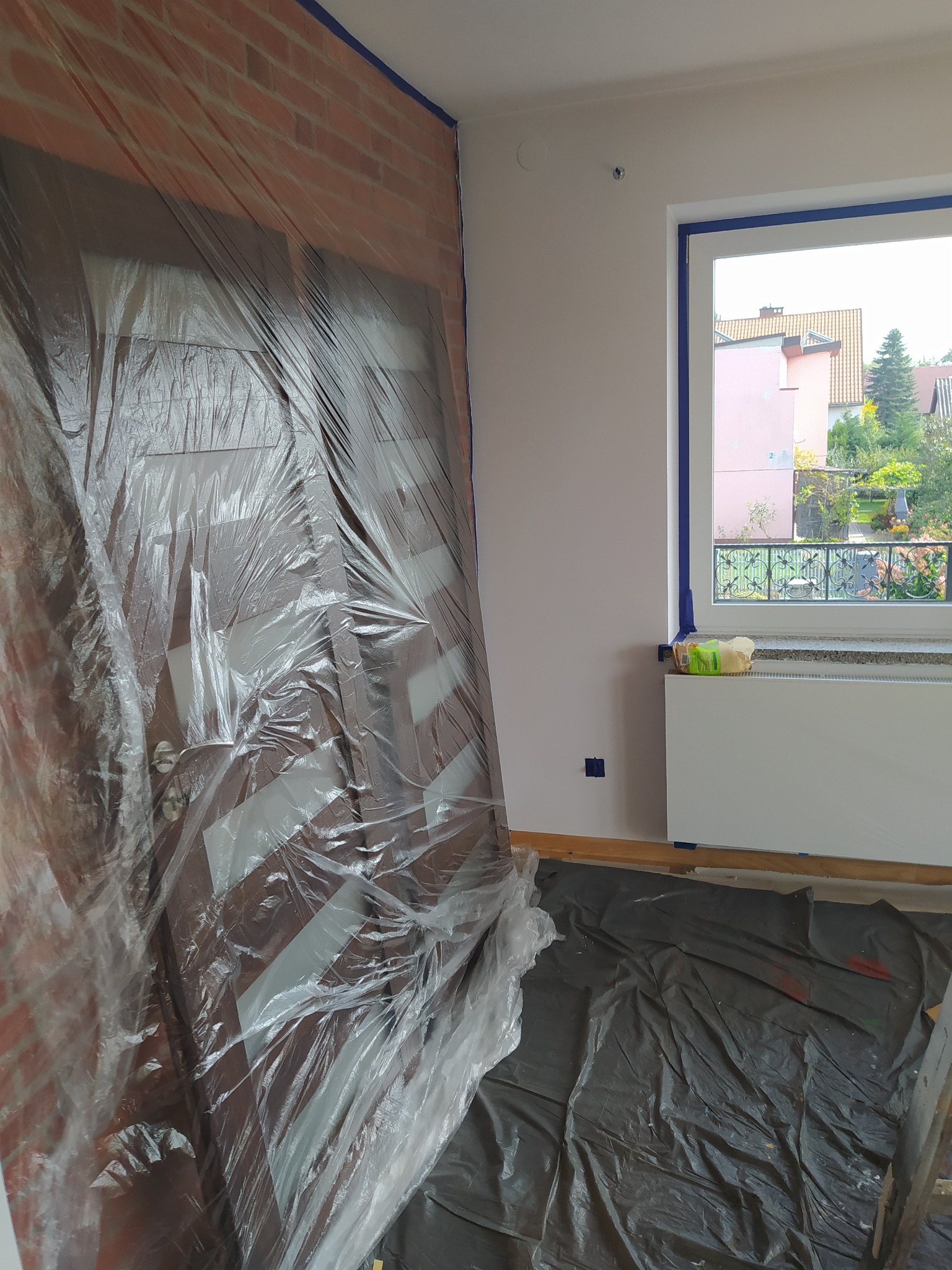 Przygotowania do prac malarskich w Oświęcimiu: Pokój zabezpieczony folią malarską, okna i framugi oklejone taśmą malarską, widoczna ceglana ściana i fragment grzejnika.