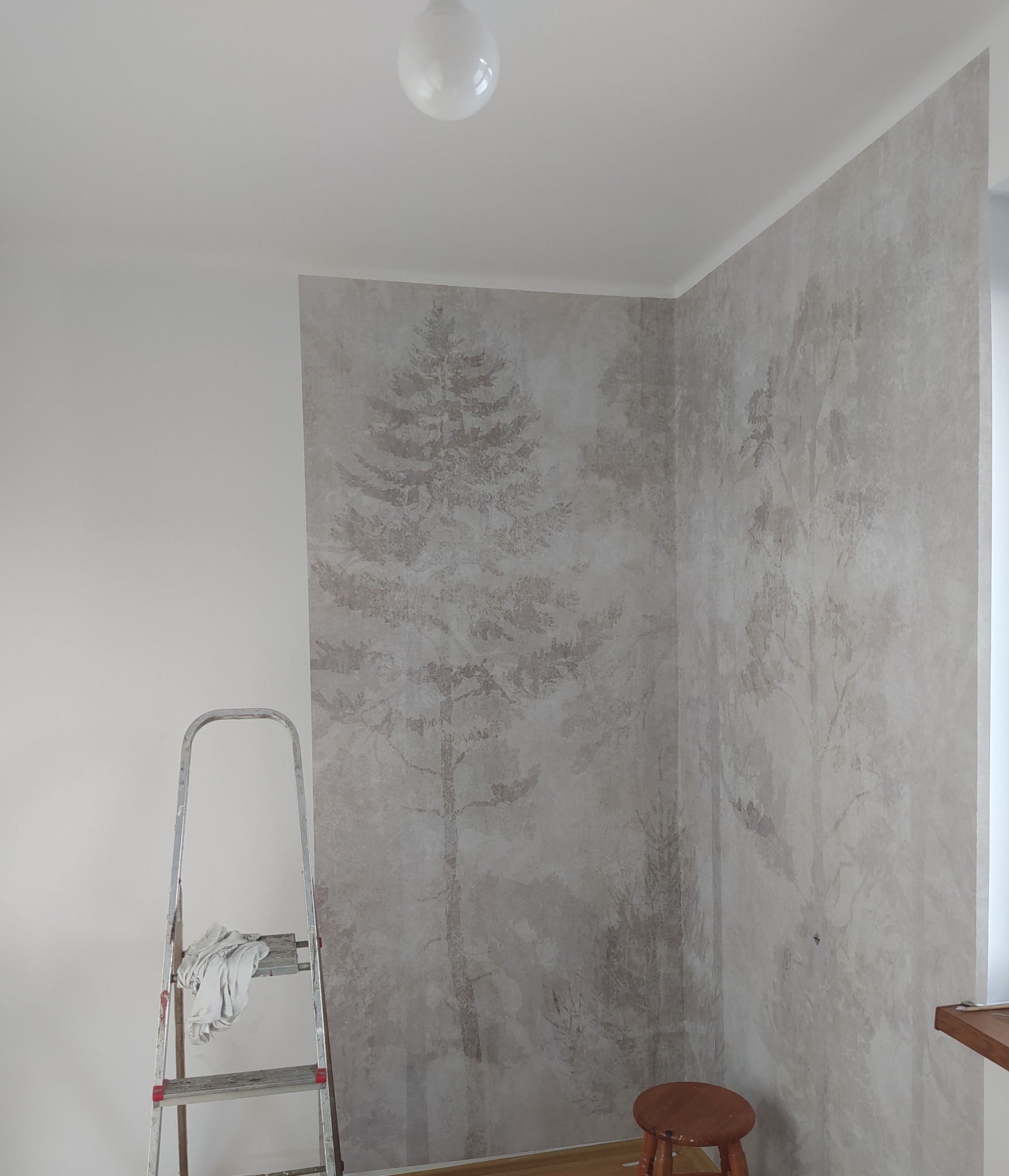 Tapeta z motywem leśnym na ścianie, drabina malarska z szmatką, drewniany stołek w rogu pokoju.