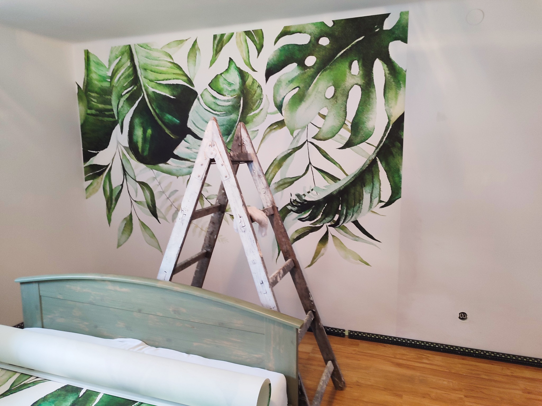 Ściana z motywem tropikalnych liści, drabina malarska oparta o ścianę, fragment łóżka w kolorze miętowym z rolką tapety w tle.