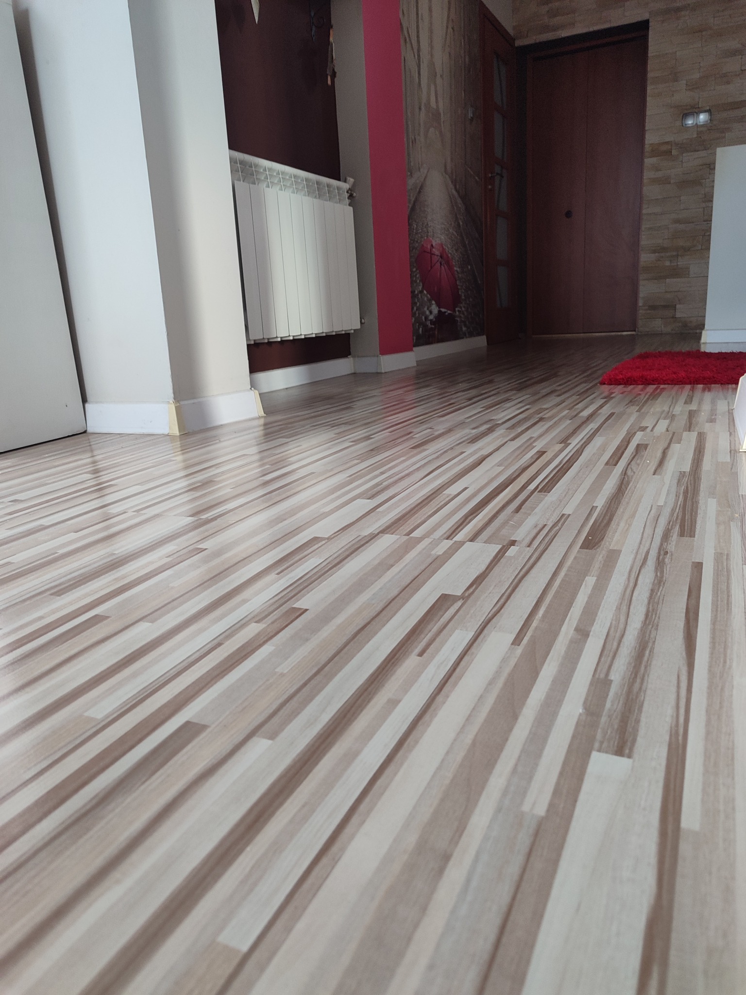 Ułożone panele podłogowe z wzorem drewna, widok z niskiej perspektywy, na podłodze czerwony dywanik, w tle ściana z tapetą z motywem uliczki i czerwonym parasolem.