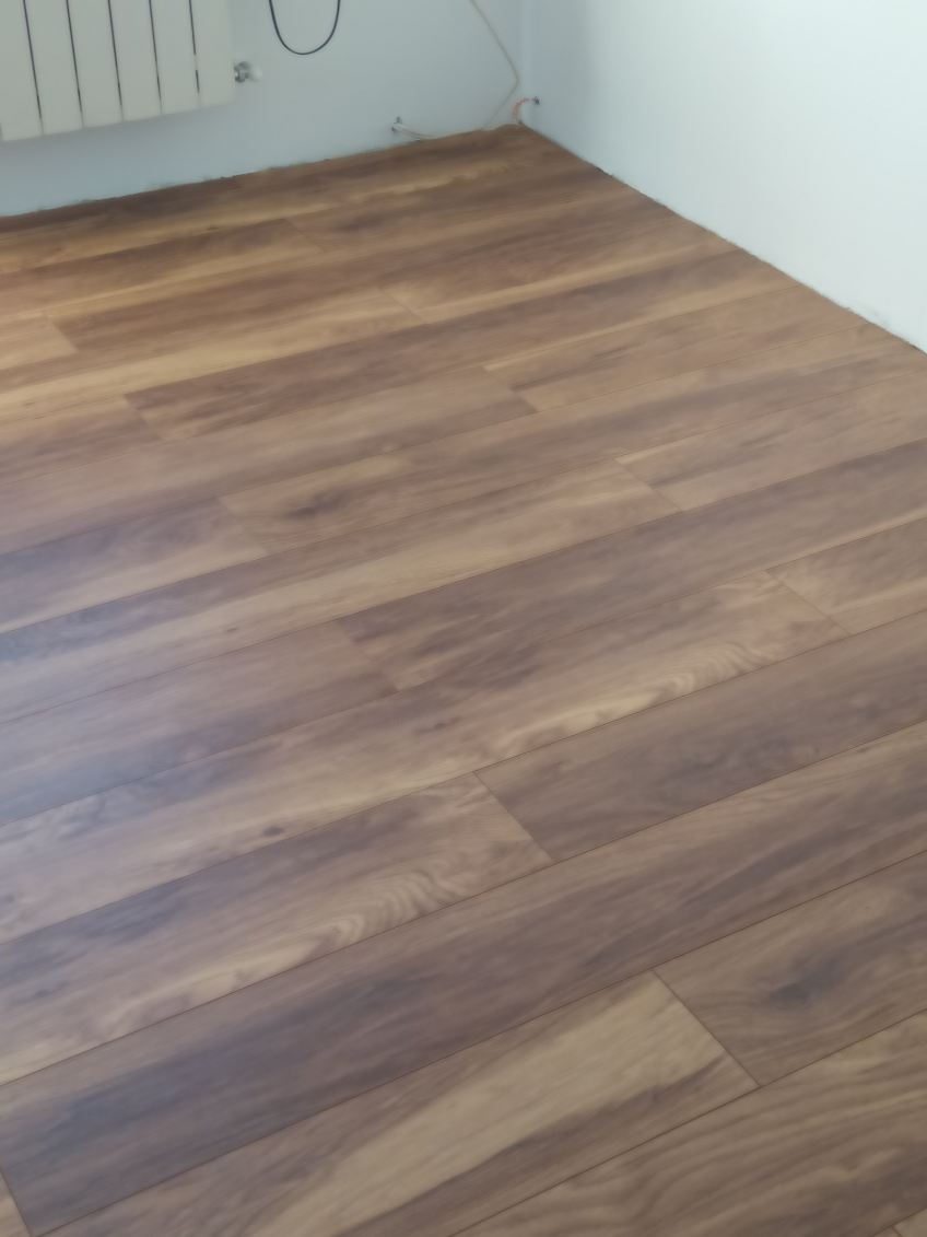 Ułożone panele podłogowe w odcieniach brązu, widoczne przy ścianie z grzejnikiem, kable wystają ze ściany.