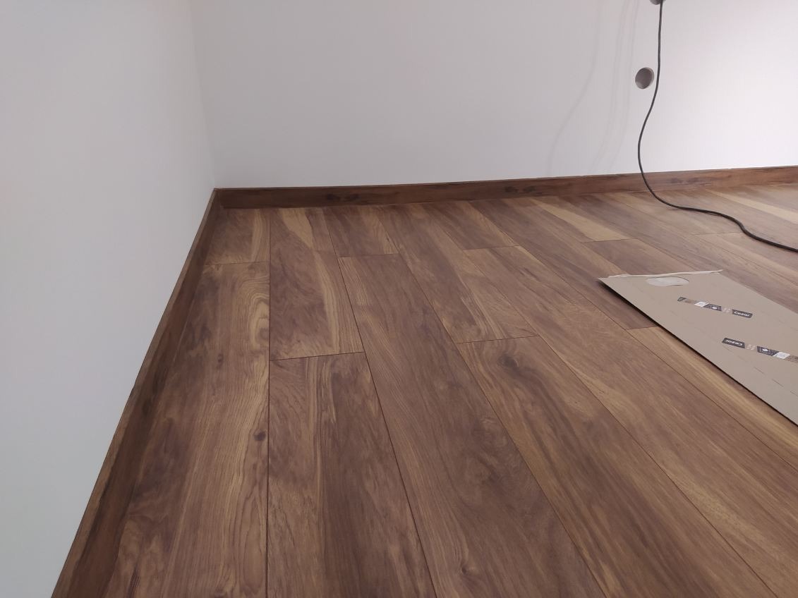 Ułożone panele podłogowe w odcieniach brązu z widoczną listwą przypodłogową, karton po panelach leżący na podłodze, gniazdko elektryczne na ścianie.